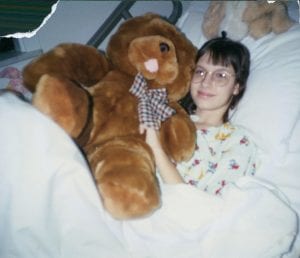 Carolyn, a childhood cancer survivor, holds a teddy bear in a hospital bed. 