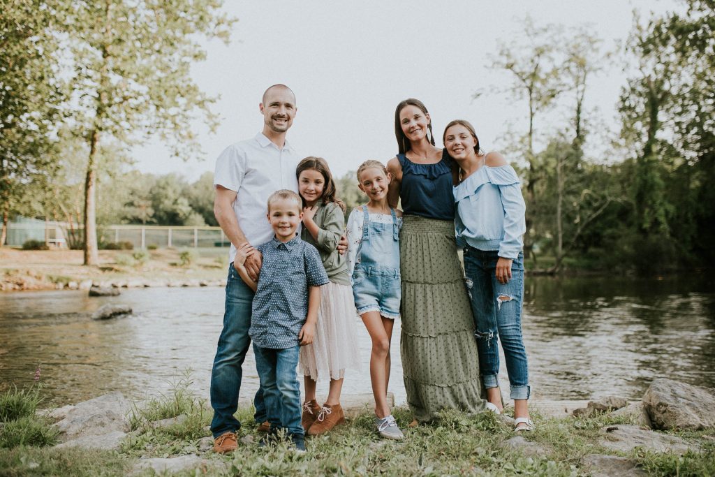Joanna Dennstaedt, melanoma survivor and founder of Radiant Hope, and her family pose and smile in front of a shining river with trees in the background