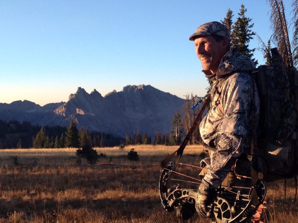 Greg Muhonen wears camoflauge attire and carries a bow with the Rocky Mountains in the background