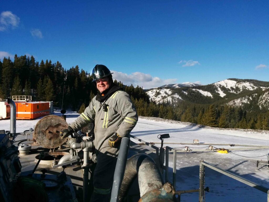 Al Drinkwalter, a melanoma survivor, wears a heavy coat and black hard hat while working near a gas pipeline with the Canadian Rockies in the background. 