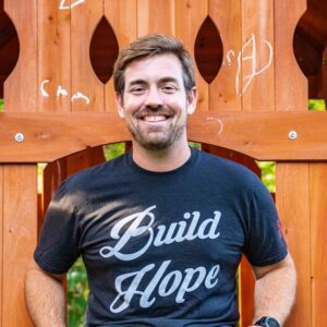 Eric Newman, the founder of Roc Solid Foundation, smiles and sits on top of a playset wearing a shirt that says, "Build Hope."