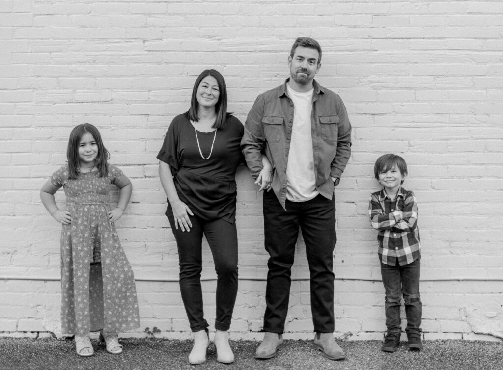 Eric Newman poses with his wife and two kids with a white brick wall as a background. 