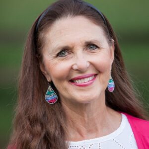 Lynn Eib, author and cancer survivor, smiles while wearing a white blouse and pink sweater and turquoise earrings. 